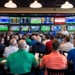 Um bar esportivo lotado com pessoas assistindo ansiosamente a várias telas mostrando diversos eventos esportivos enquanto seguram bilhetes de aposta.