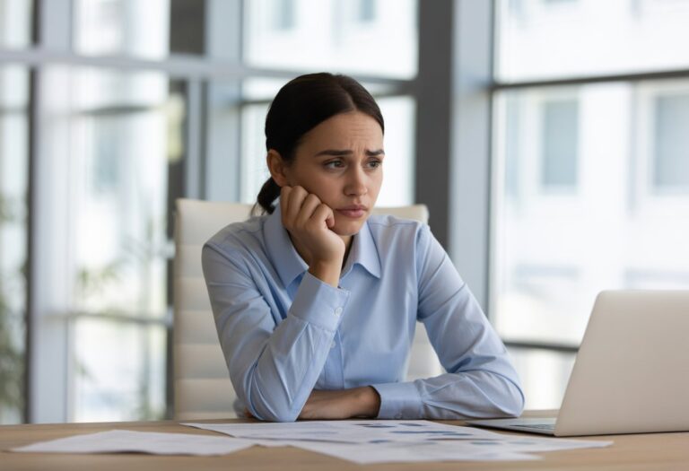 Mulher jovem pensativa sentada em uma mesa de escritório moderno, com expressão de dúvida e papéis ao redor.