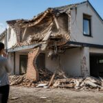 Pessoa olhando para uma casa moderna parcialmente destruída com destroços caindo ao redor.